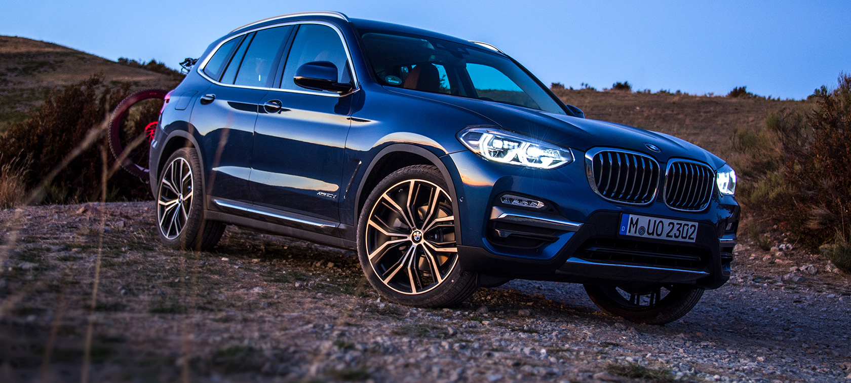 Temno model BMW X3 v naravi Parkiran temno moder BMW X3 z nosilcem in gorskim kolesom v naravi