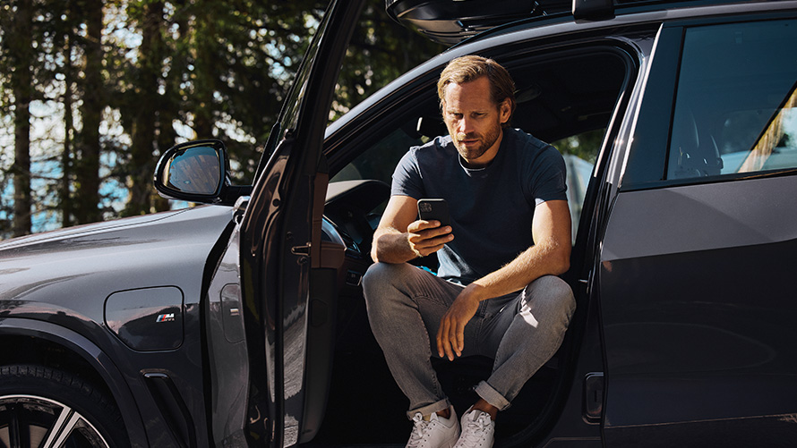 Moški sedi v BMW in gleda svoj pametni telefon Dodatki BMW za pomlad in poletje, moški sedi v BMW in gleda svoj pametni telefon