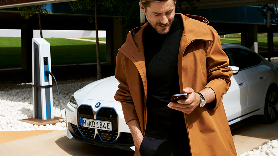 Trajanje in pogodbena obdobja BMW Service Inclusive za električna vozila BMW Service Inclusive, moški gleda v svoj pametni telefon pred BMW i4 pri polnilni postaji