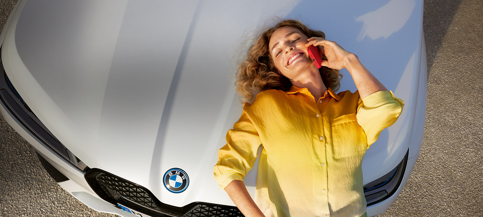 Proactive Care, pregled in vzdrževanje, sprostite se, mar nam je, ženska z rumeno srajco in pametnim telefonom v roki se naslanja na pokrov belega BMW