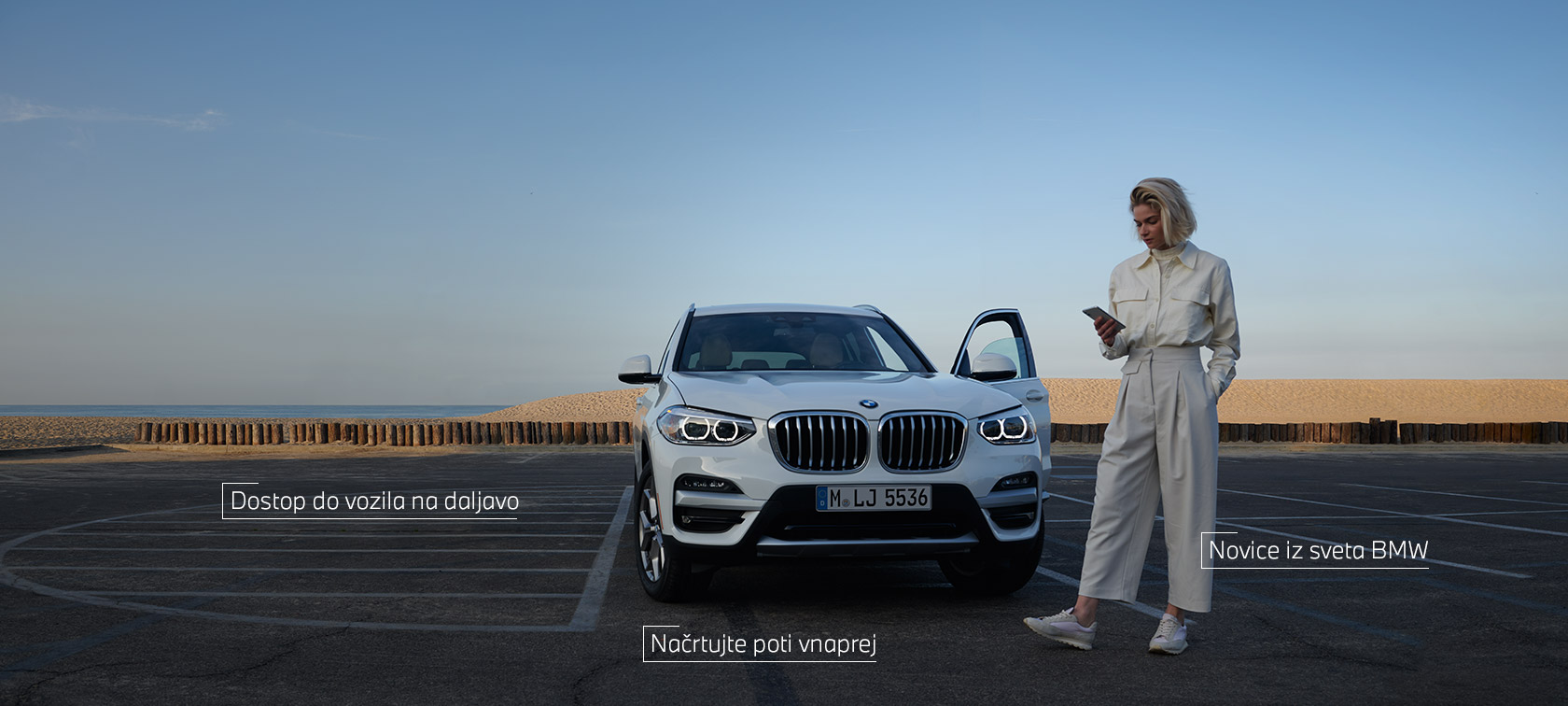 Ženska stoji pred belim BMW X5 in uporablja aplikacijo My BMW na pametnem telefonu Ženska stoji pred belim BMW X5 in uporablja aplikacijo My BMW na pametnem telefonu