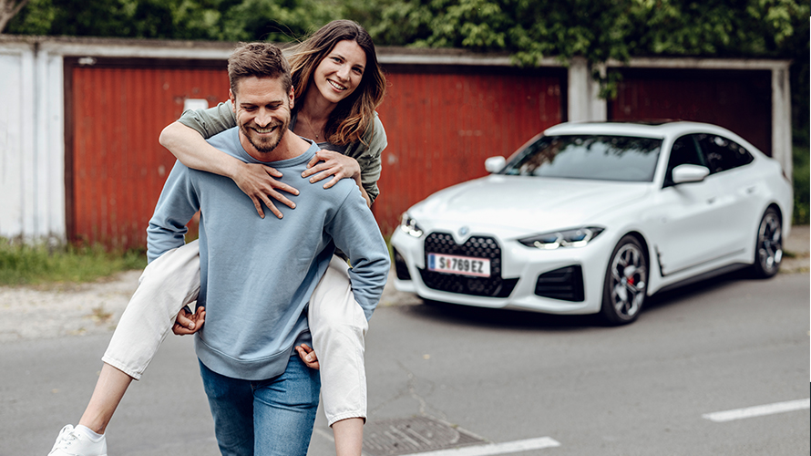 Nasmejani moški odhaja od BMW i4 in na hrbtu nosi nasmejano žensko. Nasmejani moški odhaja od BMW i4 in na hrbtu nosi nasmejano žensko.