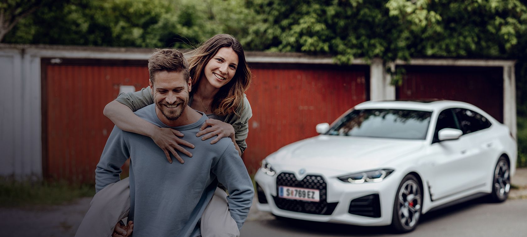 Nasmejani moški odhaja od BMW i4 in na hrbtu nosi nasmejano žensko. Nasmejani moški odhaja od BMW i4 in na hrbtu nosi nasmejano žensko.