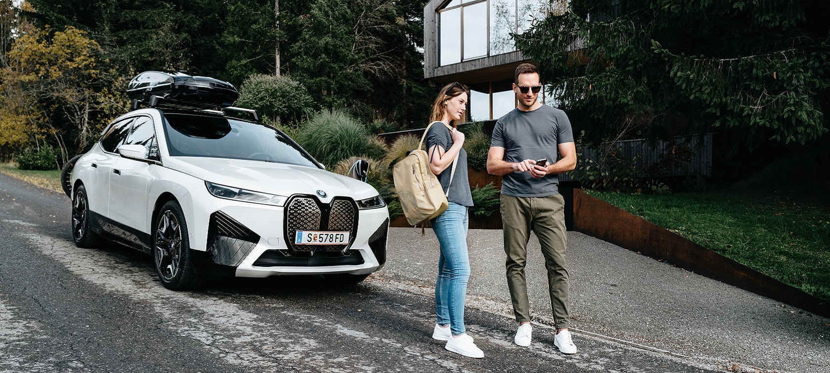 Moški s telefonom in ženska z nahrbtnikom stojita pred BMW iX. Moški s telefonom in ženska z nahrbtnikom stojita pred BMW iX.