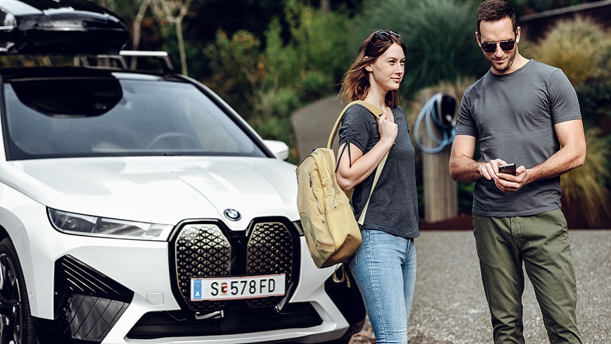 Moški s telefonom in ženska z nahrbtnikom stojita pred BMW iX. Moški s telefonom in ženska z nahrbtnikom stojita pred BMW iX.